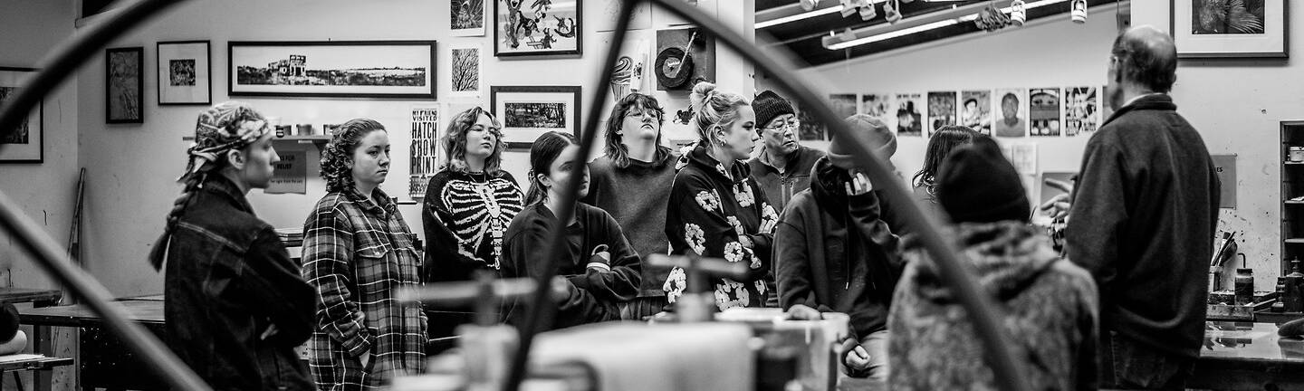 A group of individuals gathers in a workshop space, attentively listening to a speaker, with artwork displayed on the walls and industrial lighting overhead.